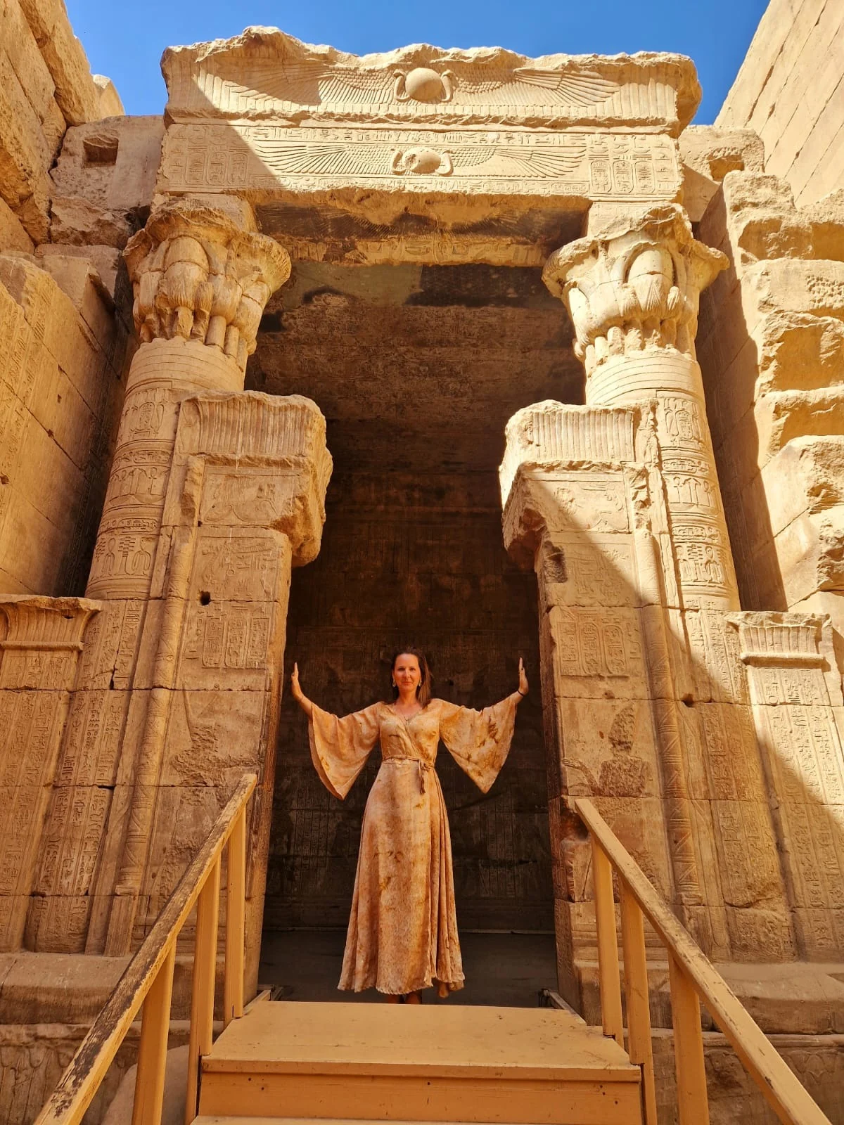 Loukia Papa with her arms raised in an Egyptian temple doorway, beneath the carved winged sun